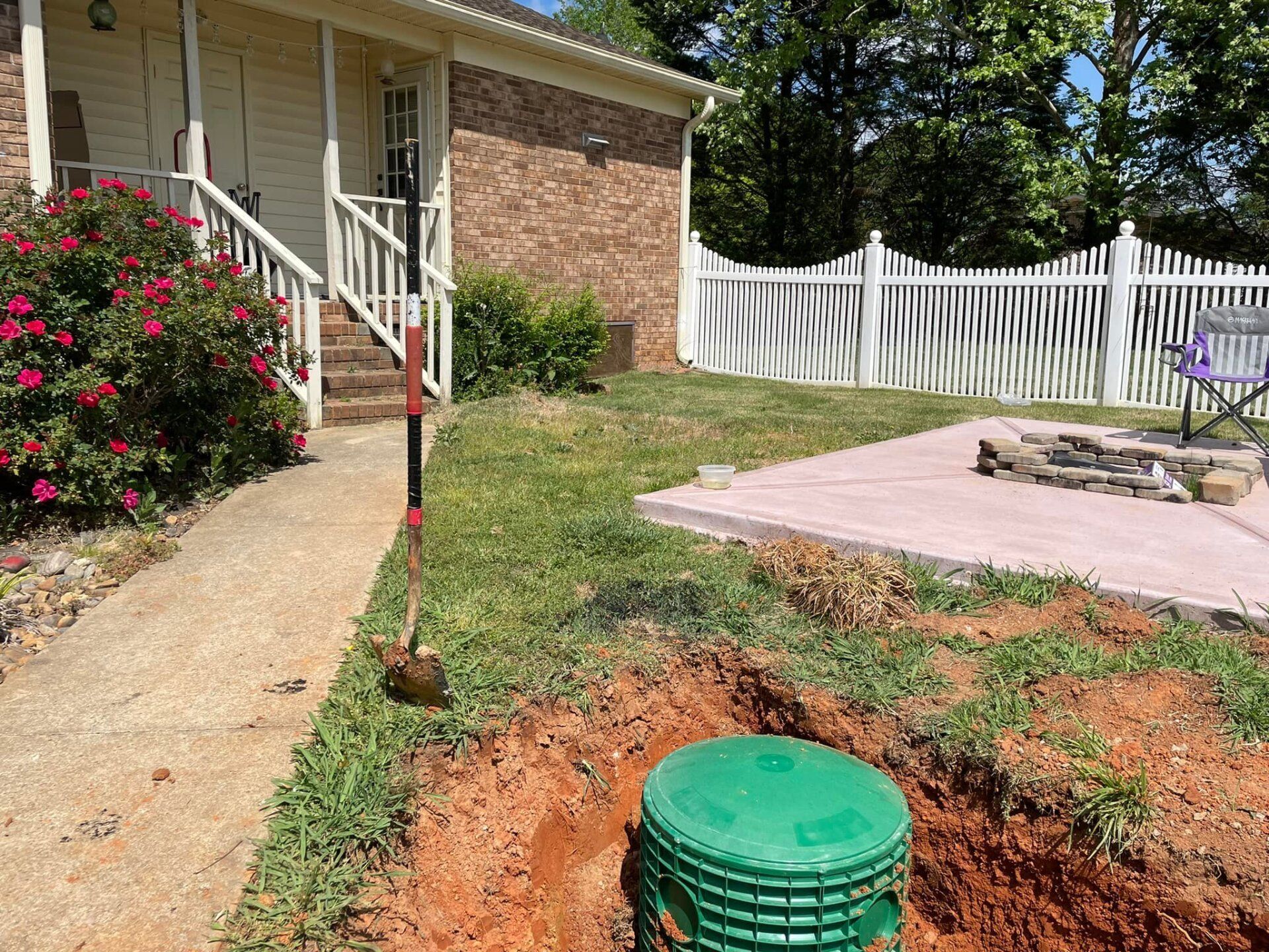 Green septic tank in excavated dirt next to a patio, house, and white fence.