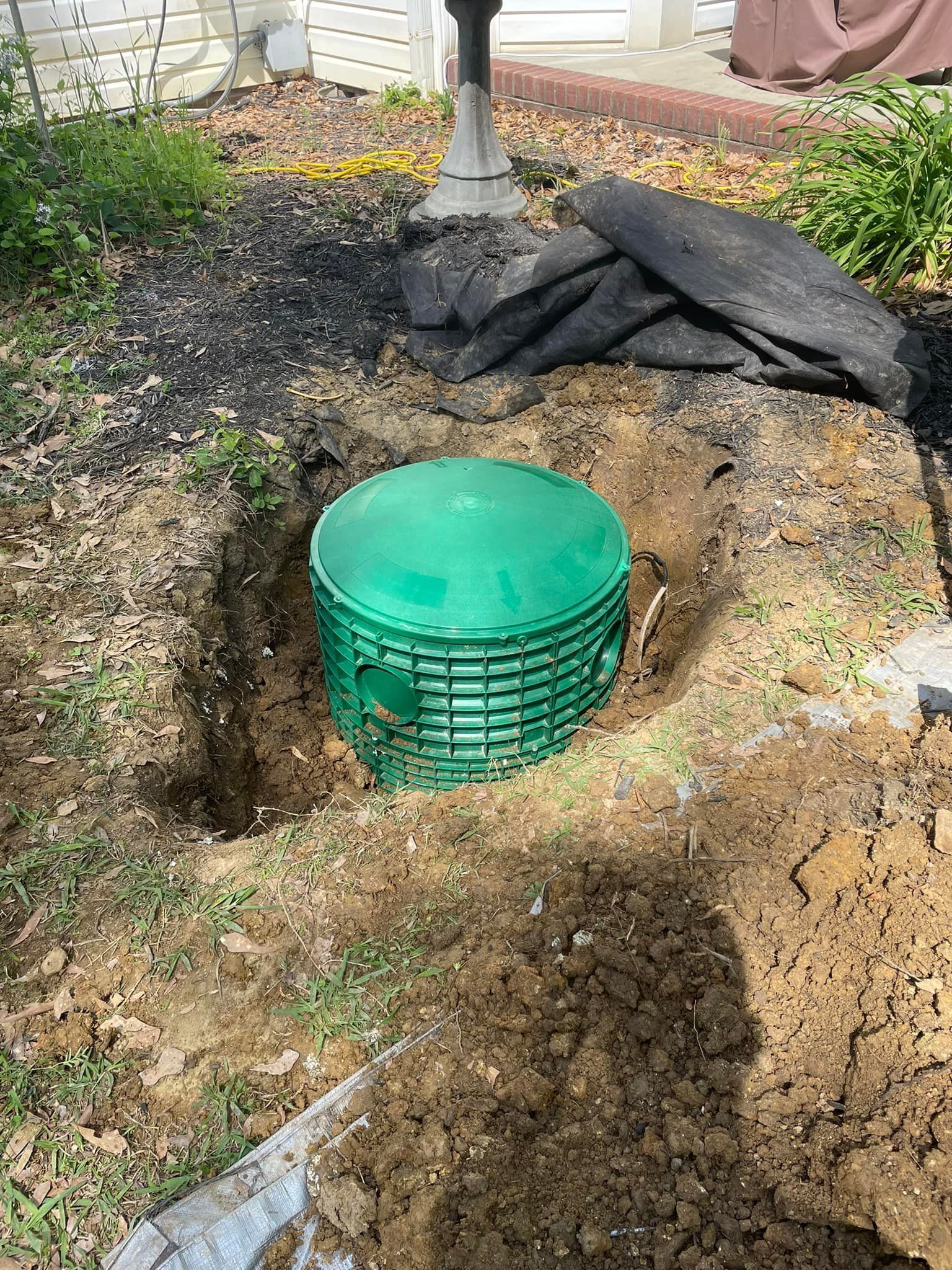 Green septic tank with a round lid is installed in a dirt pit. Black mulch and a driveway are in the background.