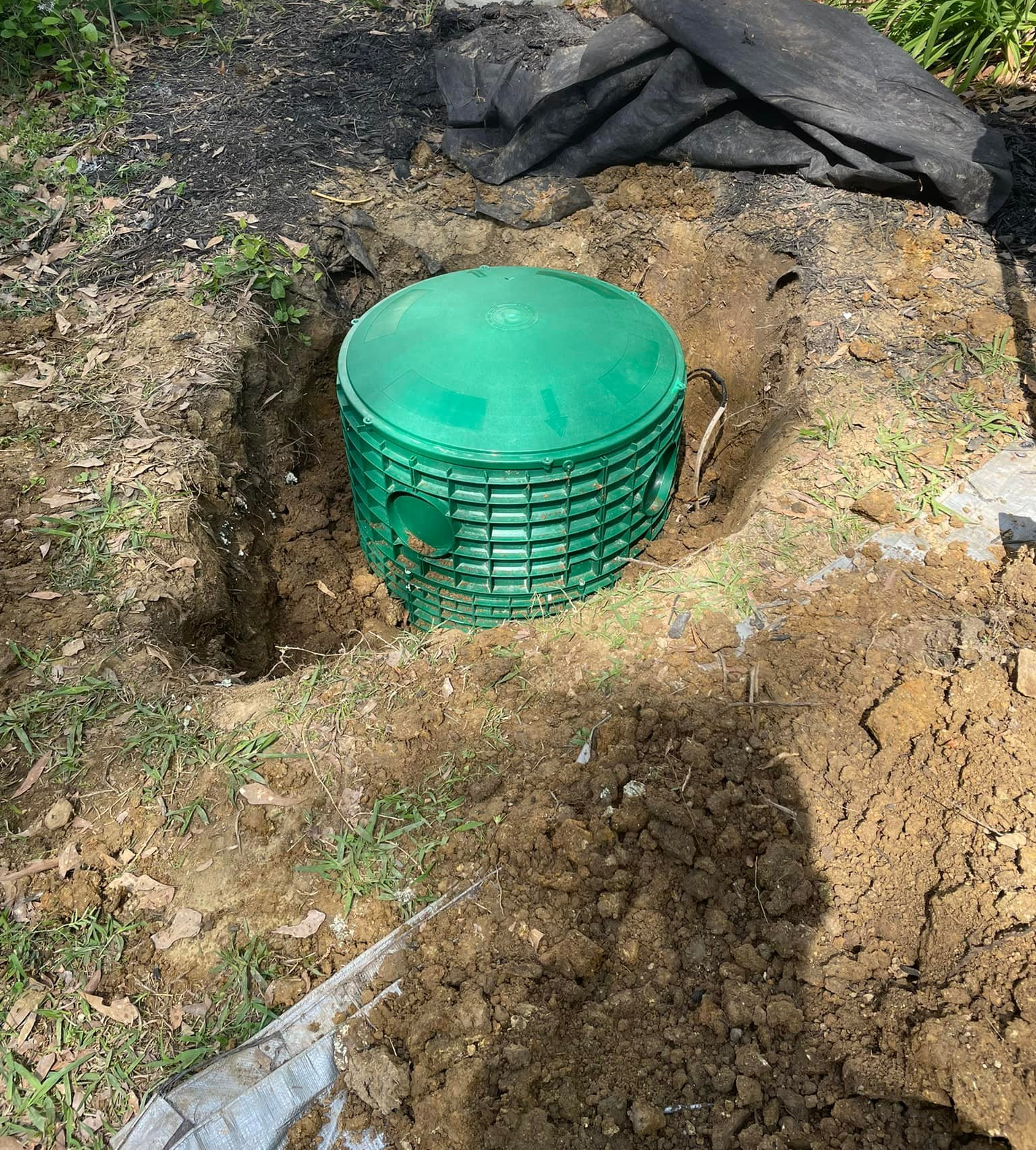 Green septic tank in an excavated hole in a yard; dirt and black fabric surround it.