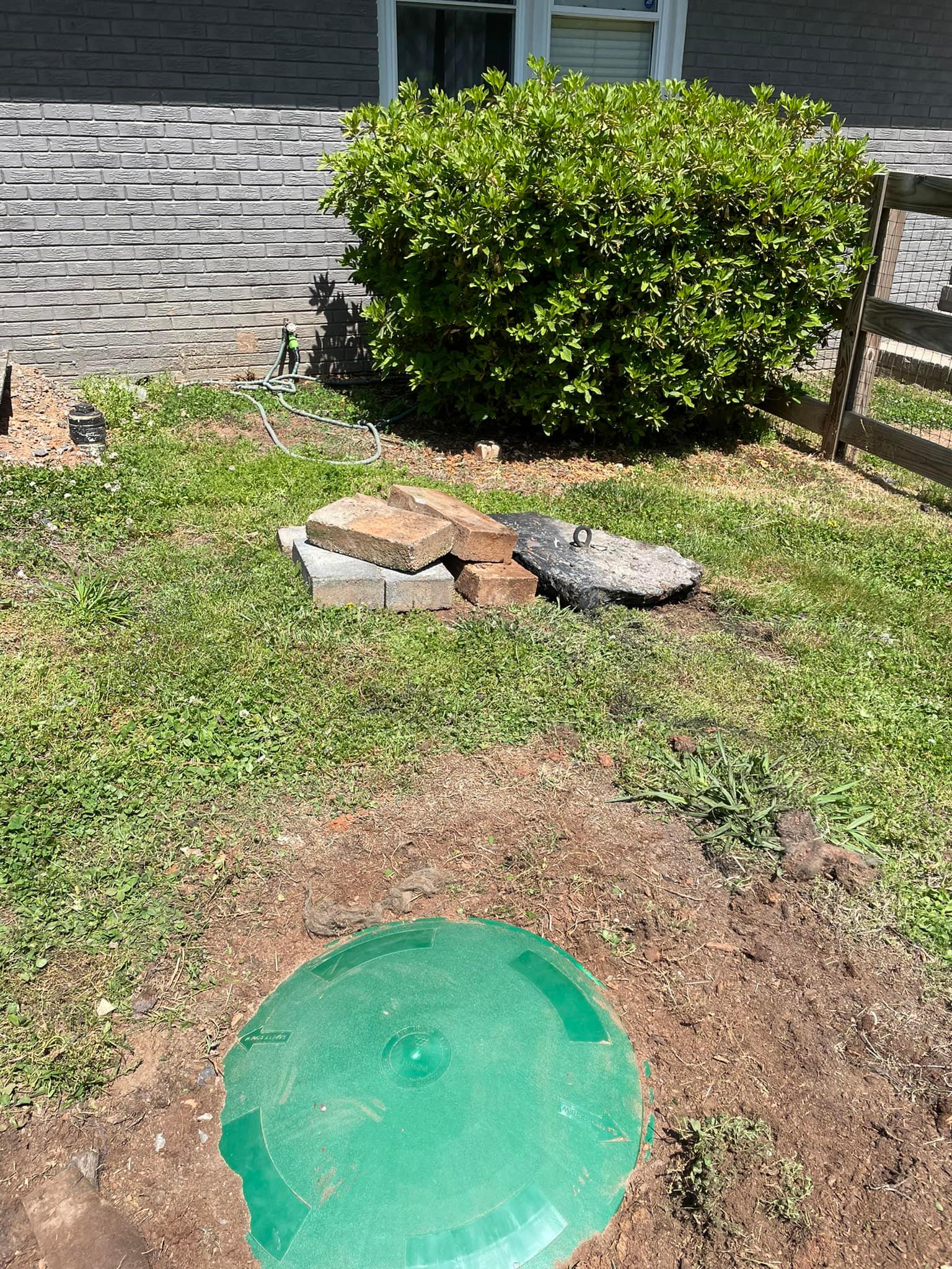 Green septic tank lid in dirt, surrounded by grass and a shrub, next to a brick stack and a house.