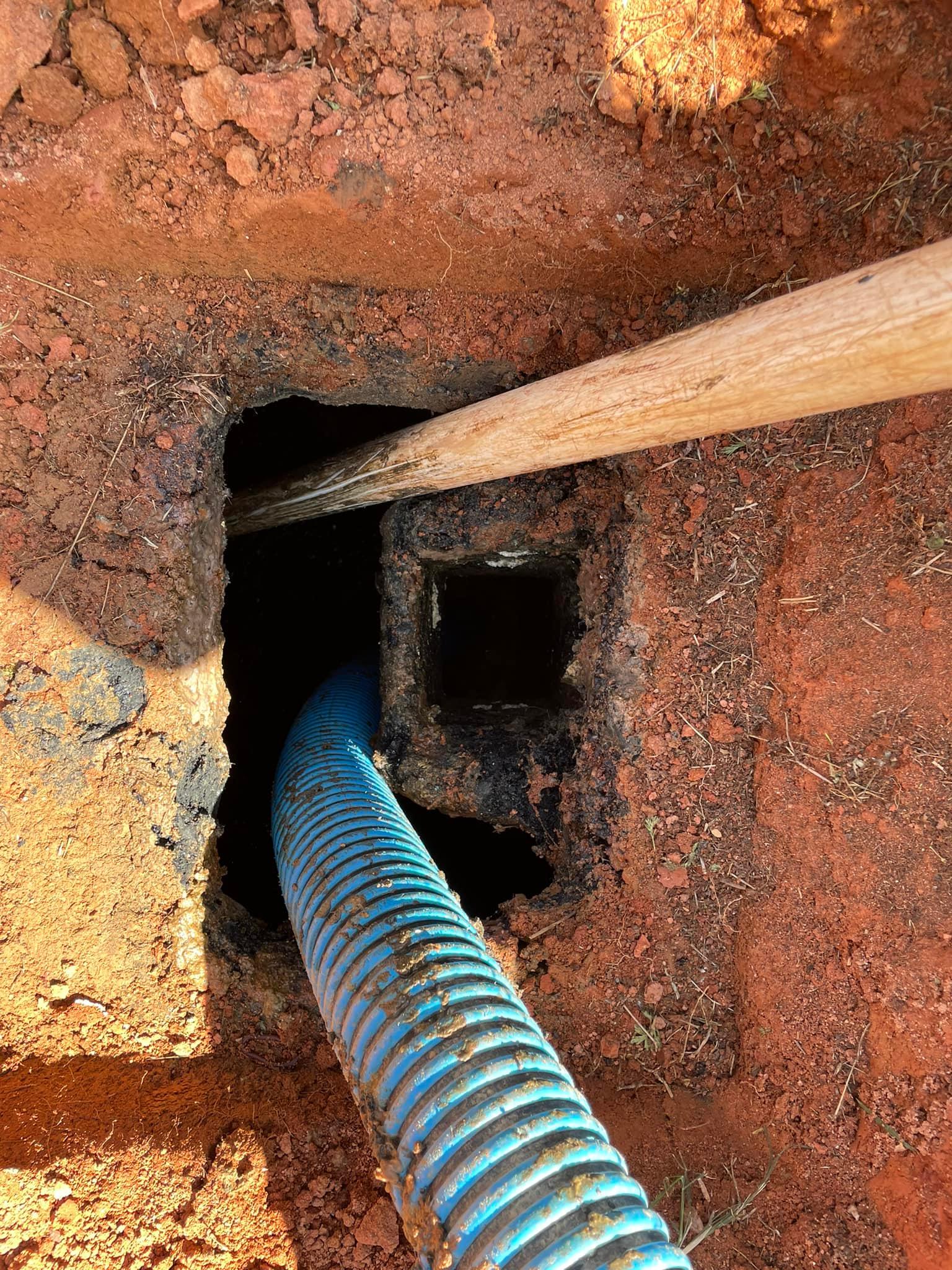 Blue corrugated pipe in an earthen trench, wooden beam across the opening.