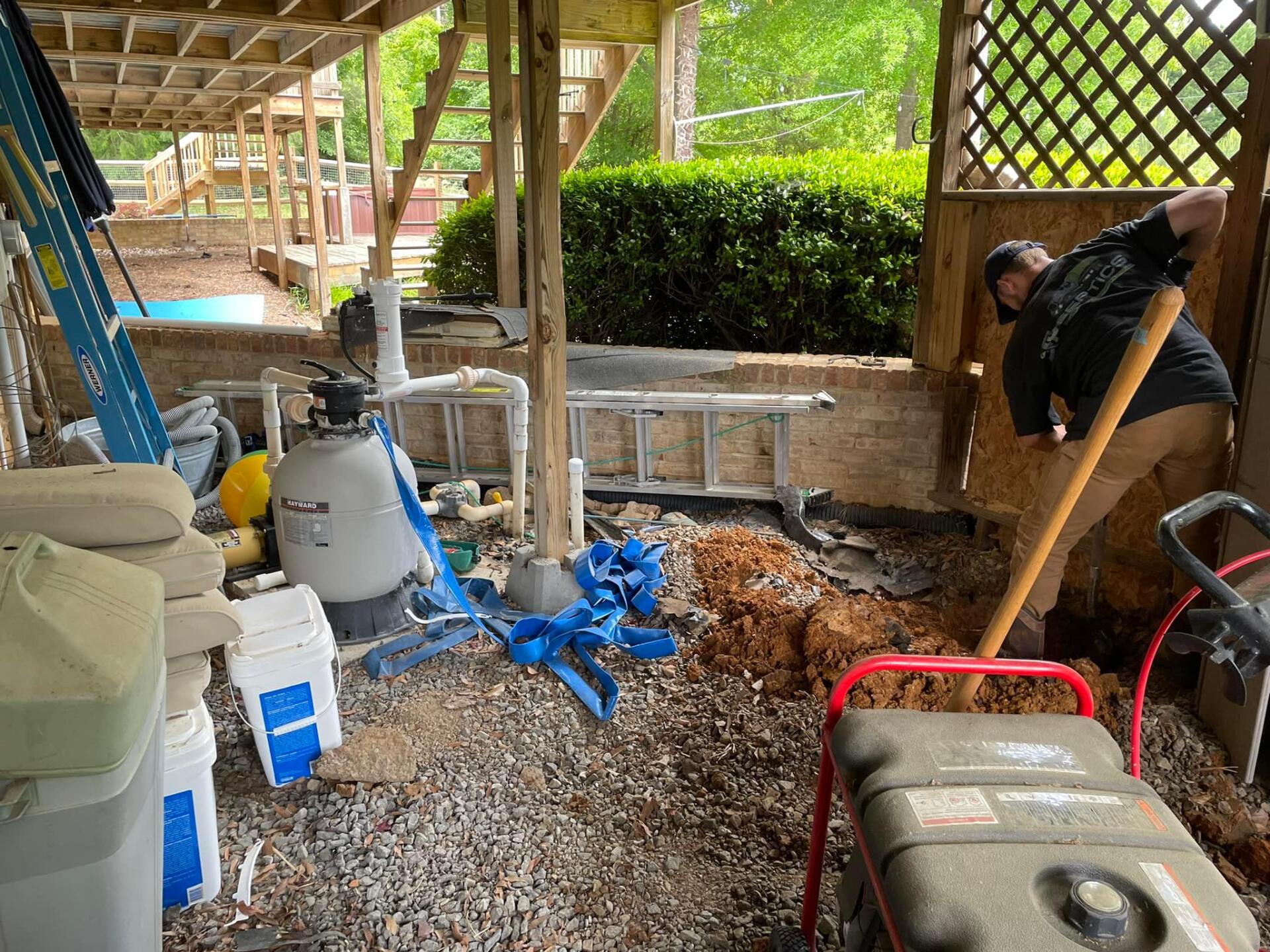 Man digging with a shovel under a wooden deck. Gravel ground, equipment and a blue tarp visible.