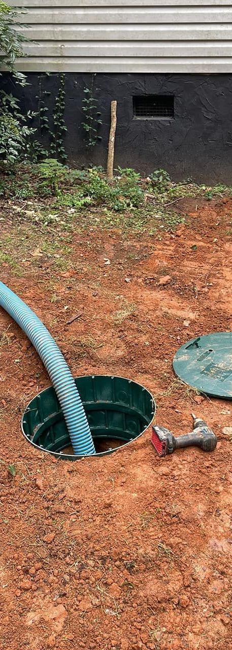 Septic tank being serviced; blue hose in open tank, red drill on ground, brown earth.