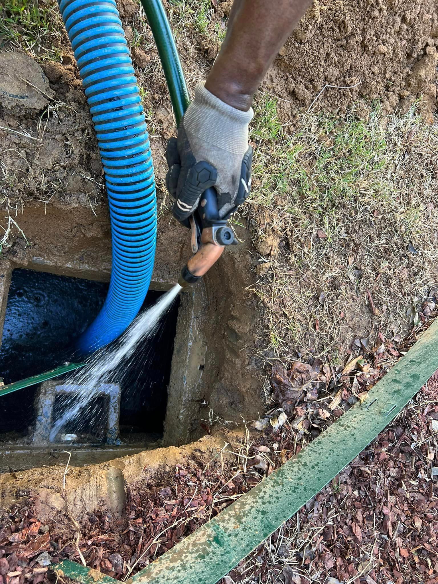 A person cleaning a septic tank. A blue hose and a green hose are connected to the tank while water sprays.
