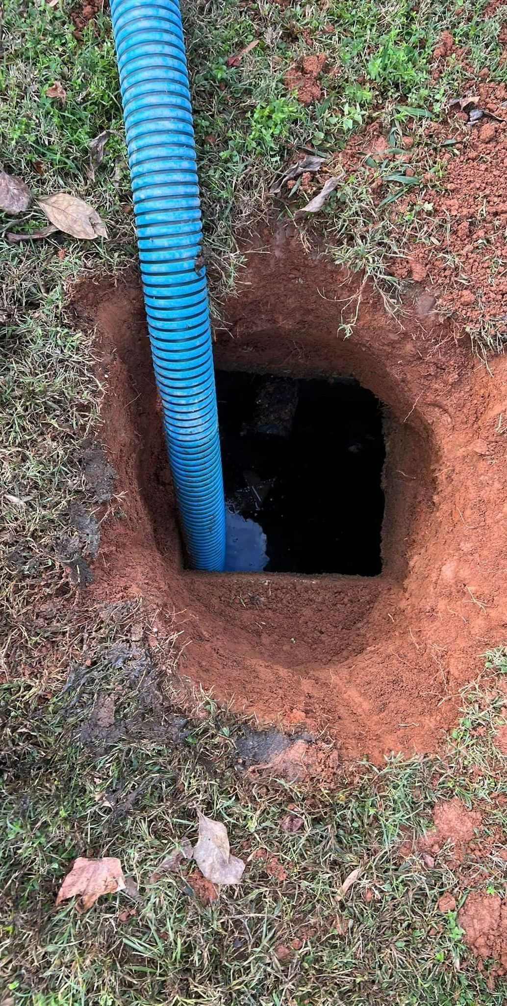 Blue corrugated pipe extends into a square hole in reddish-brown earth. Green grass surrounds the hole.