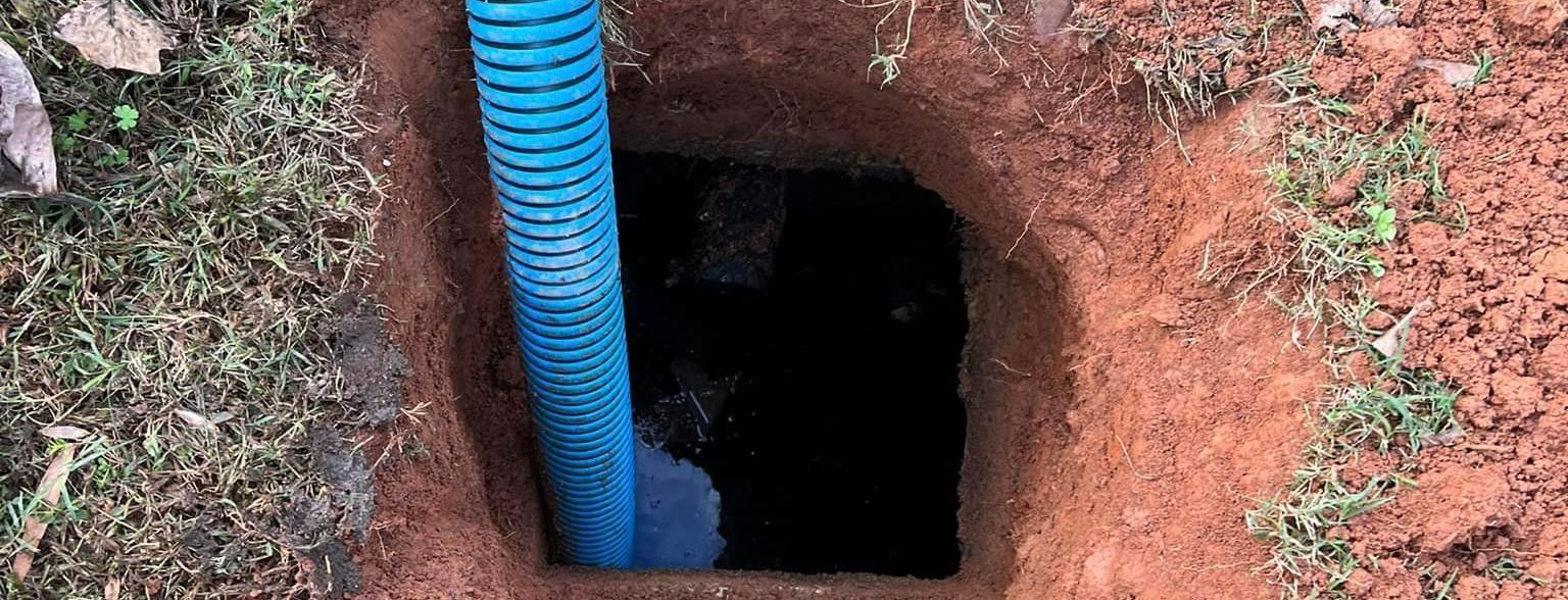 A blue corrugated pipe descending into a square hole in the reddish-brown earth, filled with dark liquid.