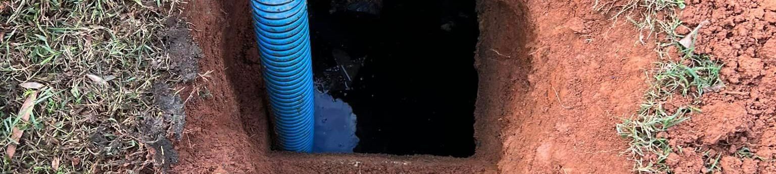 A rectangular hole dug in reddish soil with blue pipe and green grass nearby.