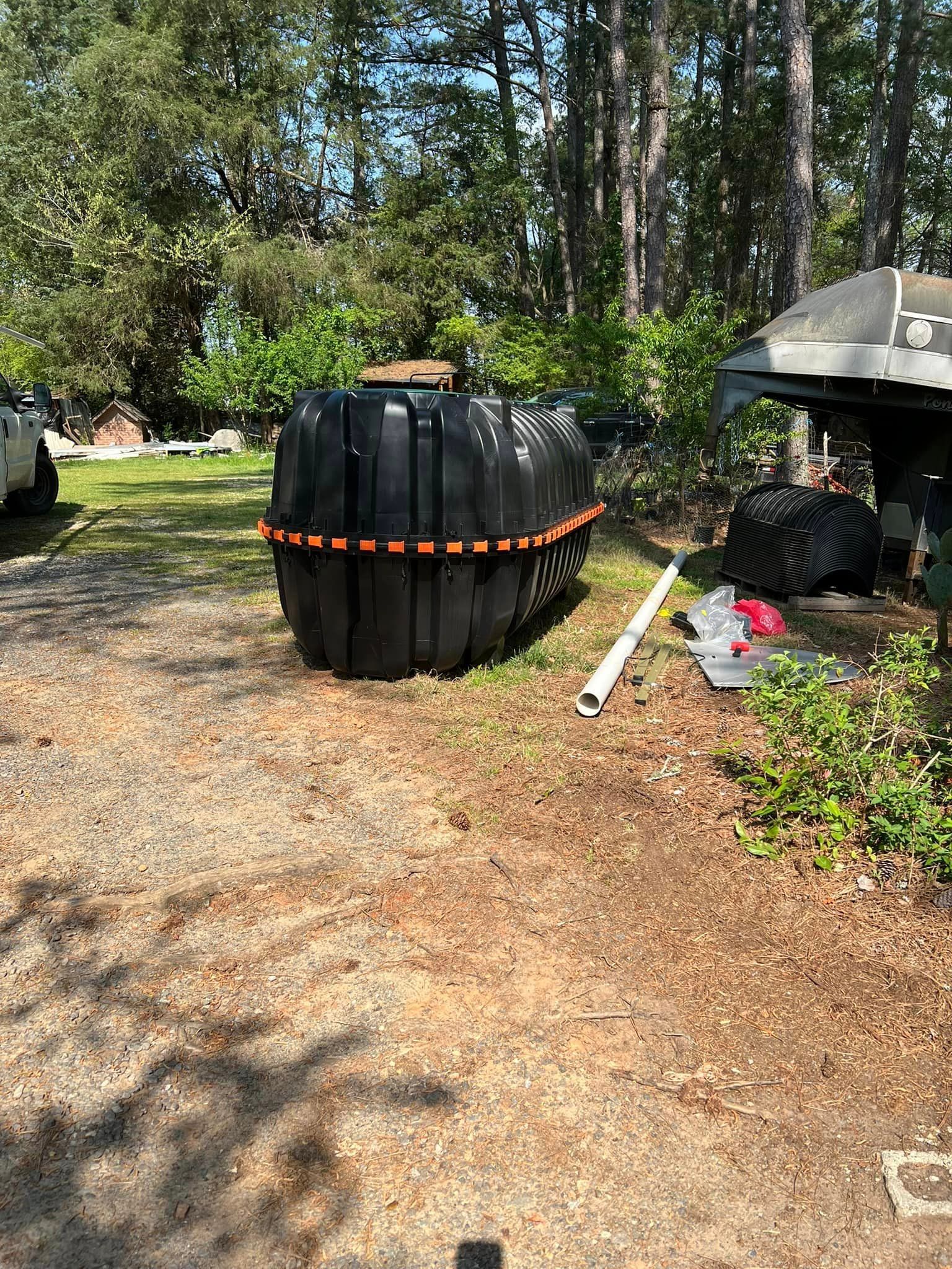 Black septic tank outdoors, surrounded by trees and gravel. White pipe and red trash bag nearby.