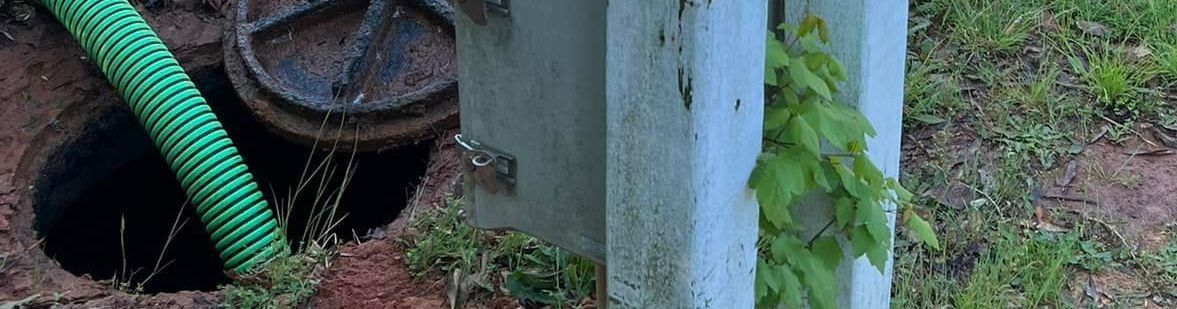 Green hose going into a dark hole; a concrete structure with green plants; grass in the background.