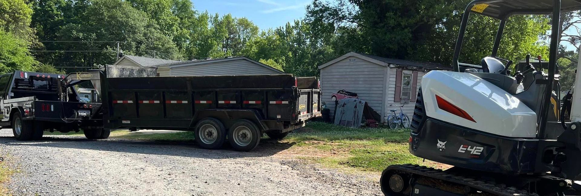 Truck towing a trailer near a small shed and an excavator. Trees and green grass are in the background.