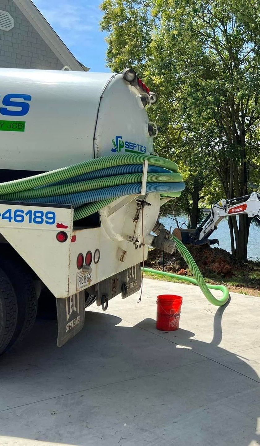 Rear view of a white septic tank truck with green hose and red bucket on a concrete driveway near trees and water.