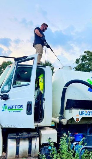Man on a septic truck spraying the tank, wearing sunglasses, blue sky.