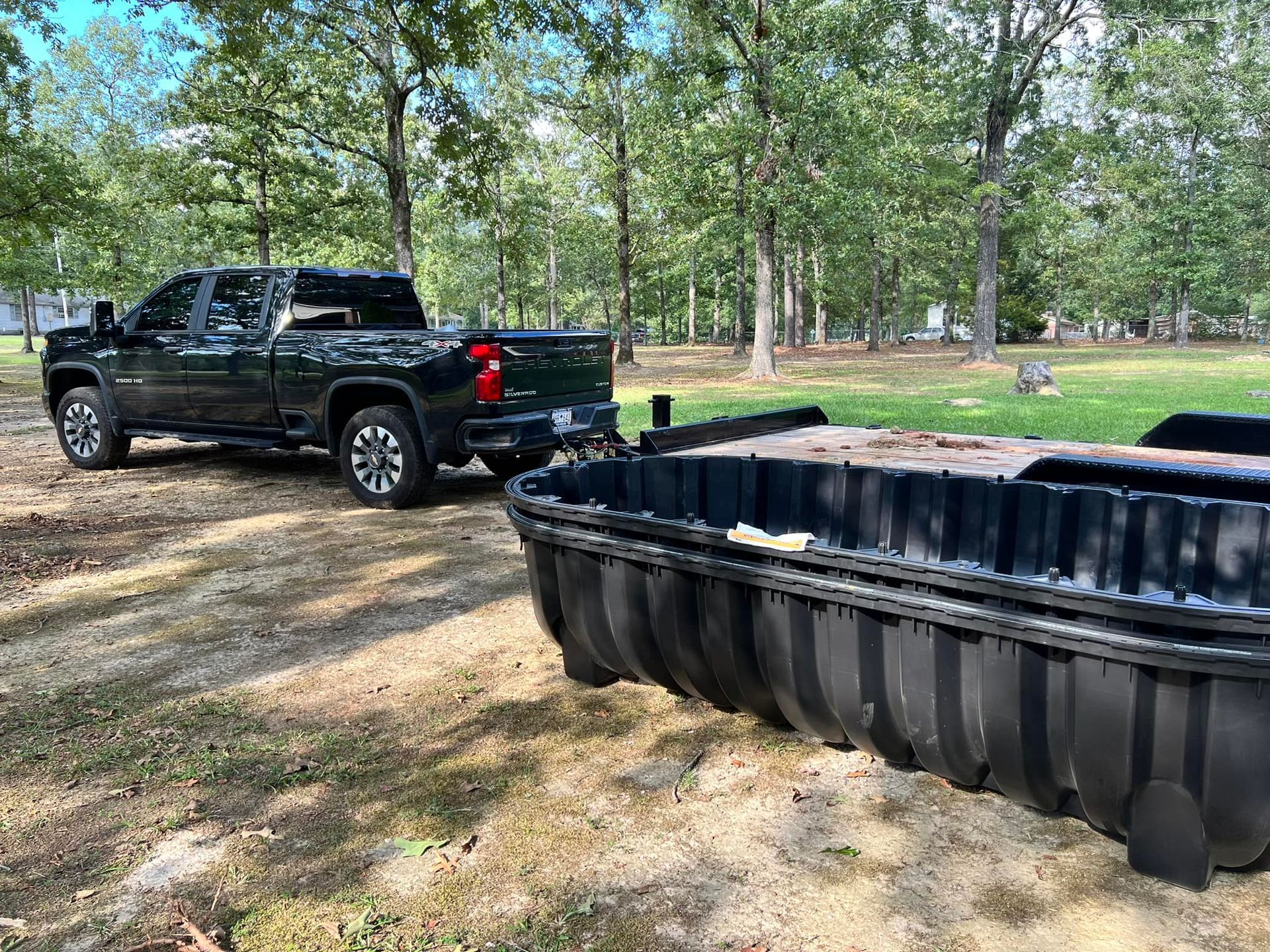 Dark pickup truck towing a black, ribbed container on a trailer, outdoors in a grassy area with trees.