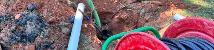Hole in the ground with green and white pipes, red and blue coils of hose, and brown dirt.