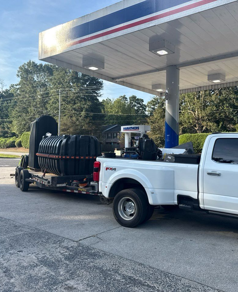 White pickup truck towing a trailer with a large black septic tank at a gas station under a canopy.