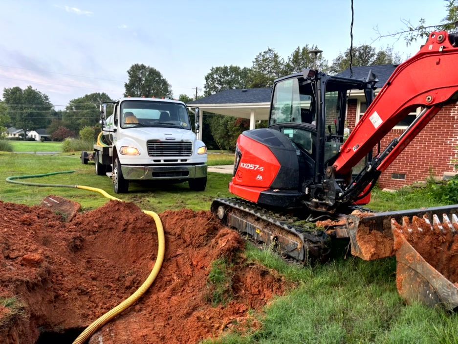 Truck and excavator working on a septic system; brown soil, green grass, house in background.