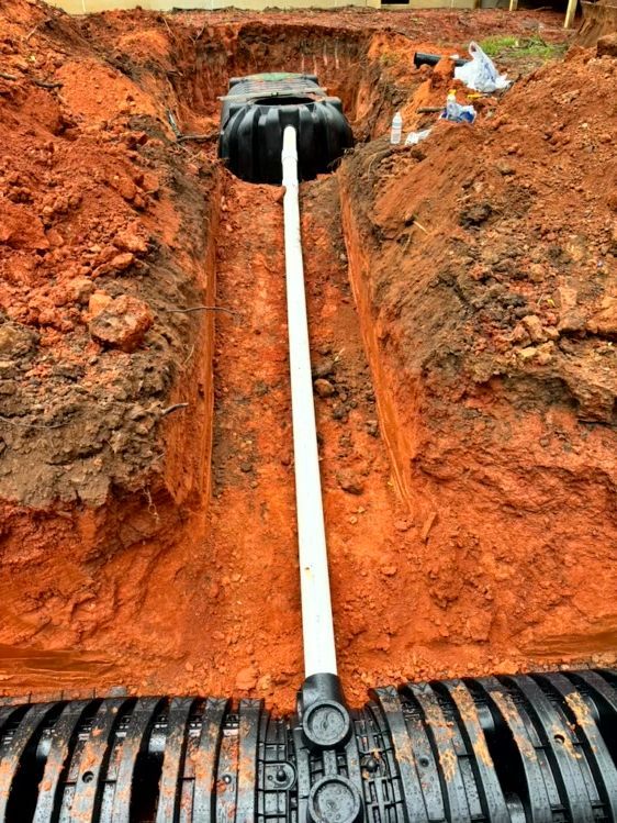 A trench with a septic tank, white pipe, and drainage system in red soil. Construction site.