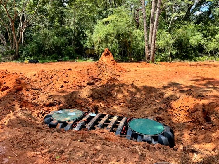 Septic tank installation in a cleared, red dirt area. The tank has two green lids and black plastic grating.