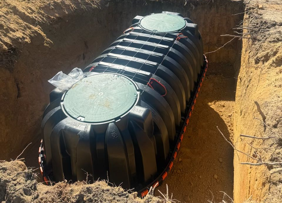 Black septic tank, orange band, in grassy yard with trees, white pipe, and shed.