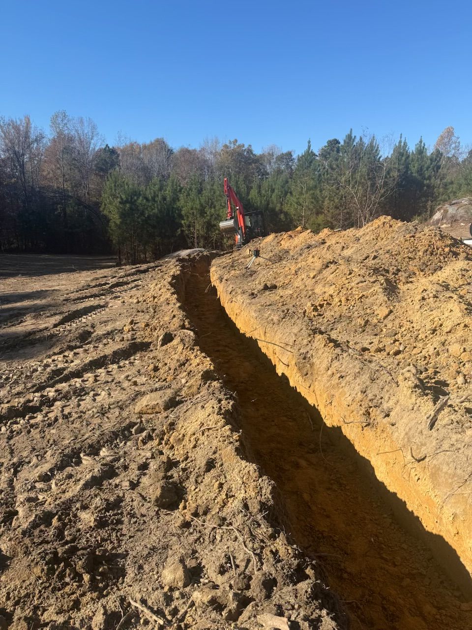 A long trench dug in brown soil, with an excavator in the distance under a blue sky.