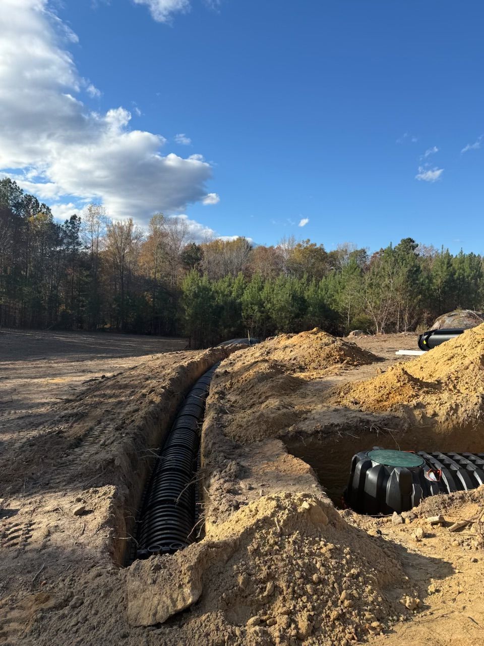An open trench with black pipes installed, surrounded by dirt and a blue sky.