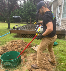 Man using a tool to work on a septic tank in a yard with a deck and grill.