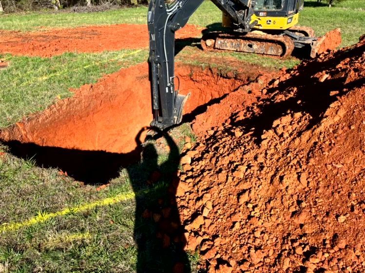 Mini excavator digging a rectangular trench in red clay. Operator's shadow visible. Green grass nearby.