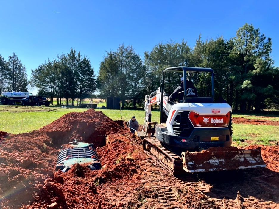 Bobcat excavator digging trench in red soil, near a septic system and trees under a blue sky.