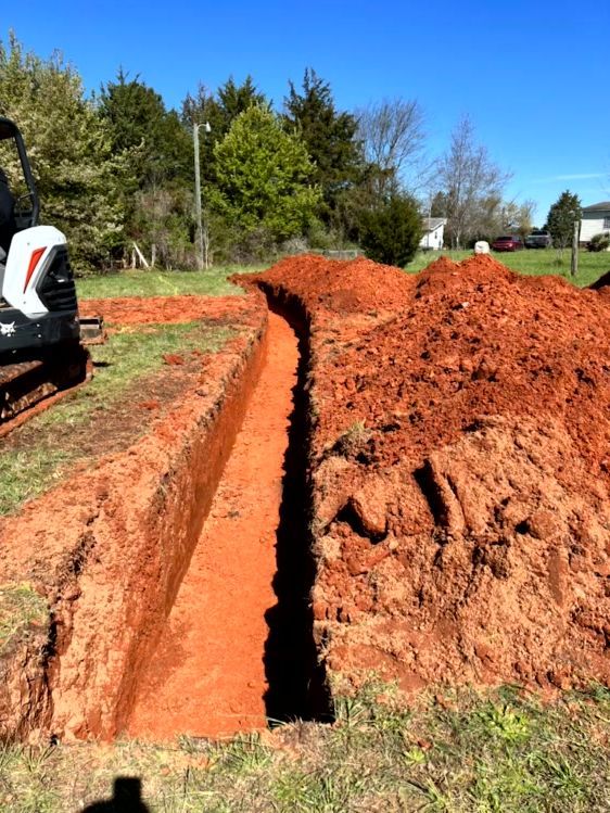 A long, narrow trench dug in reddish soil, with a small excavator visible in the background.