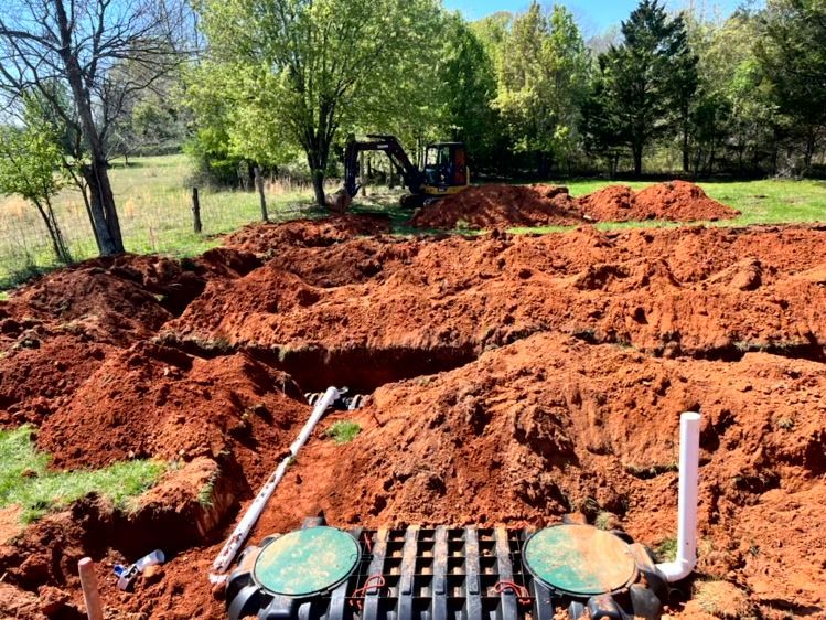 Septic system installation; brown soil, trench, excavator in background, two green lids, white pipes.