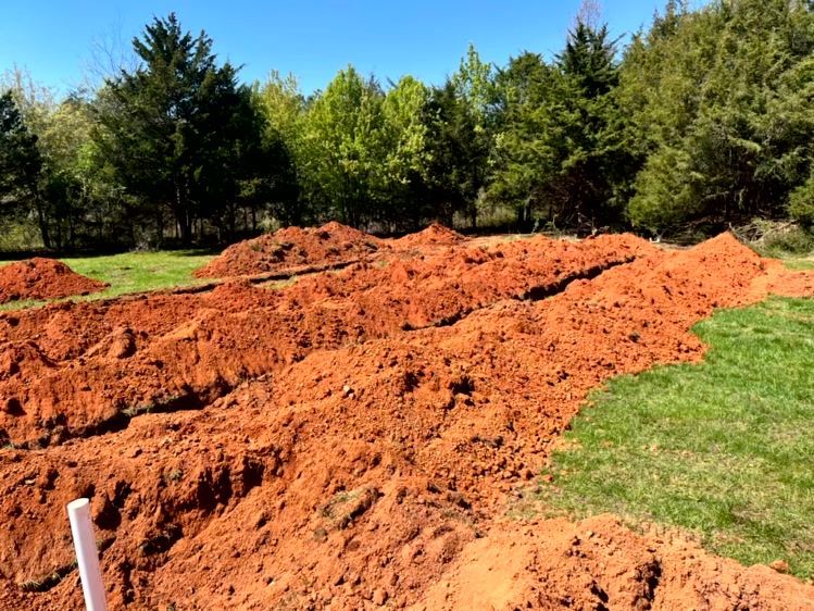 Trenches dug in red earth on a grassy area, piles of soil, trees in the background, blue sky.