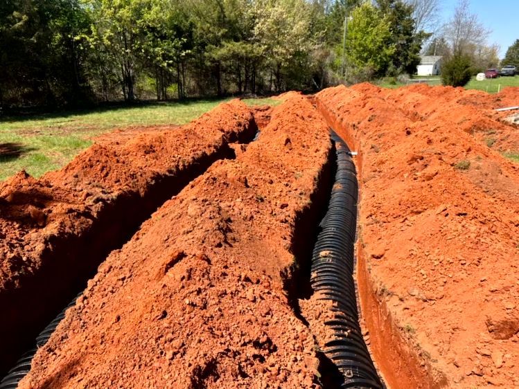 Trenches dug in red dirt with black corrugated pipe laid inside, ready for burial.