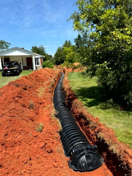Black corrugated pipe in an open trench, near a house, for drainage. Red soil and green grass surround. Sunny day.