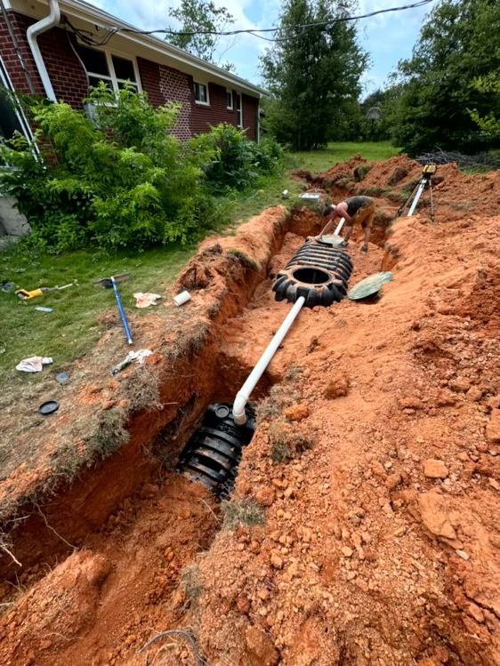 Trench with septic system components installed in reddish-brown soil near a brick house.