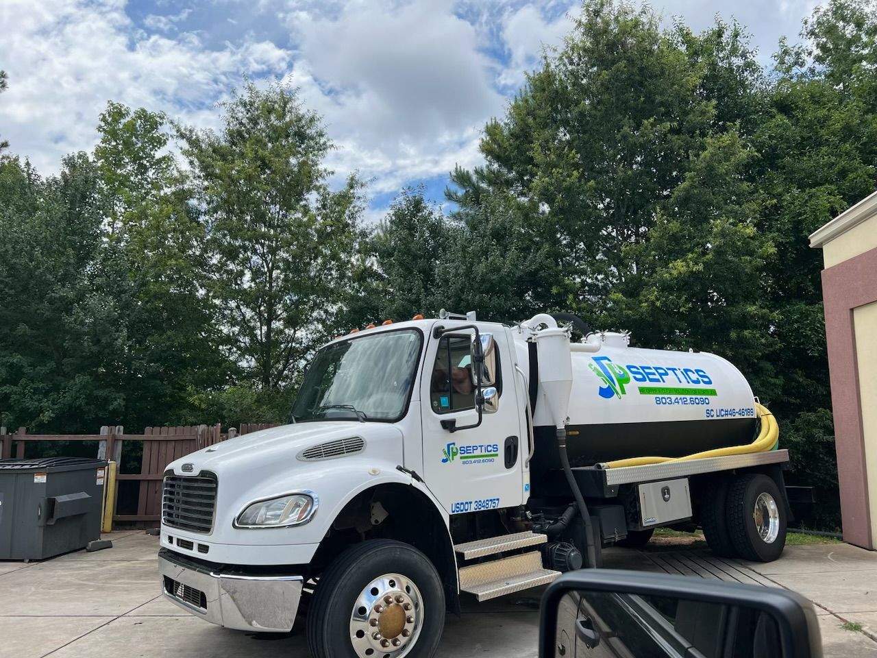 Septic service truck parked; white cab, black and white tank; green trees in background.