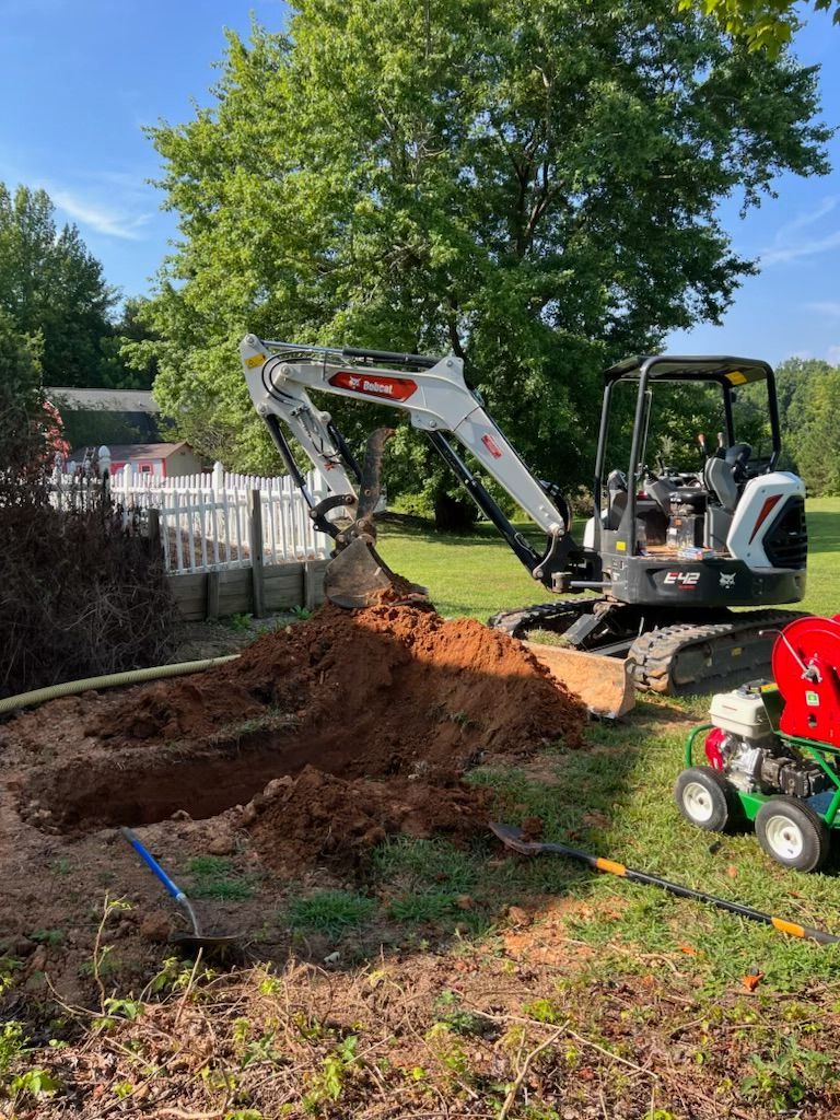 Bobcat excavator digging in a yard near a white fence and a tree.