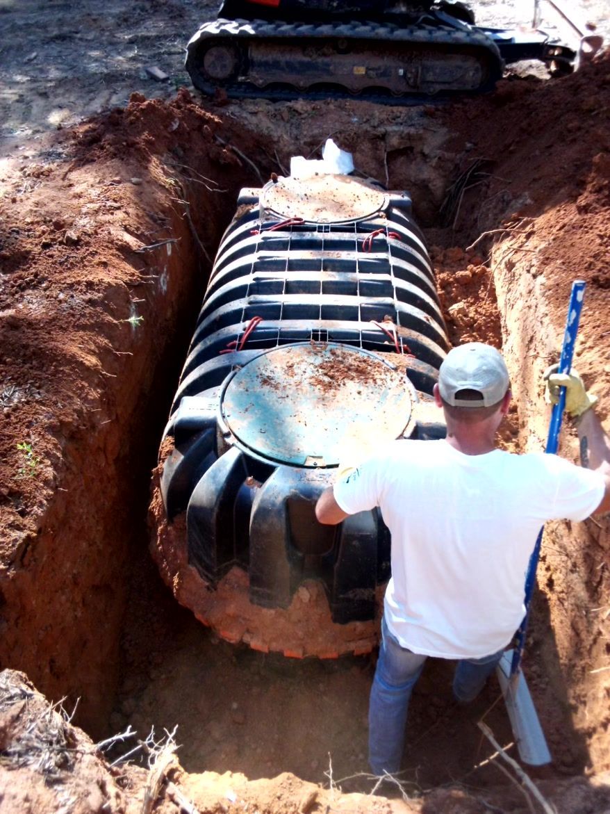 Person installing a black septic tank in a dirt trench, excavator in background.