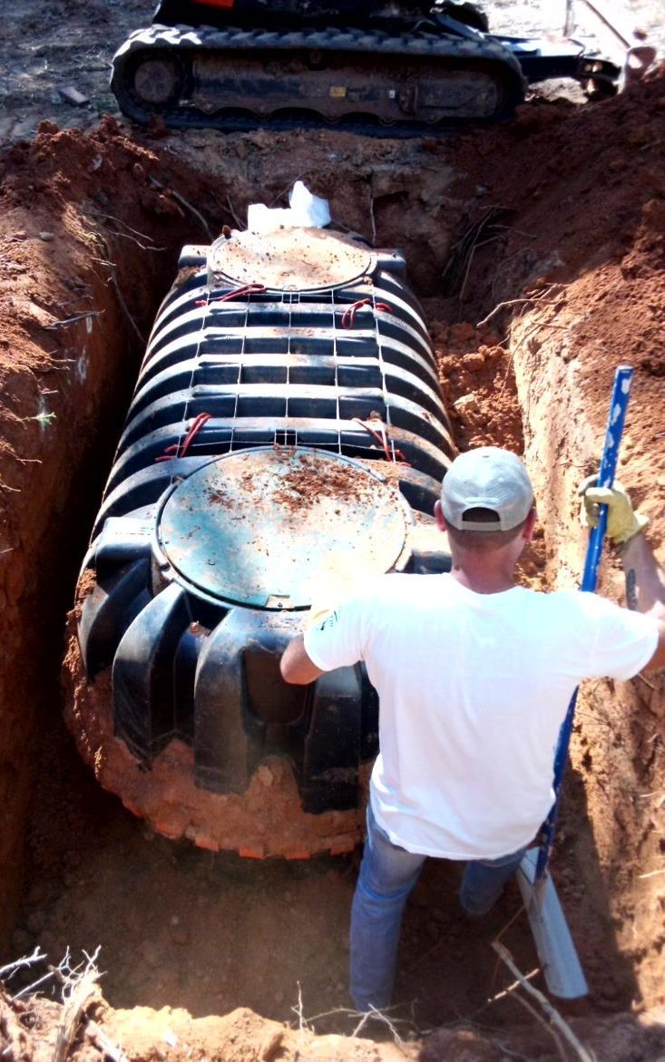 Person installing a large black septic tank in a dirt trench, excavator in background.