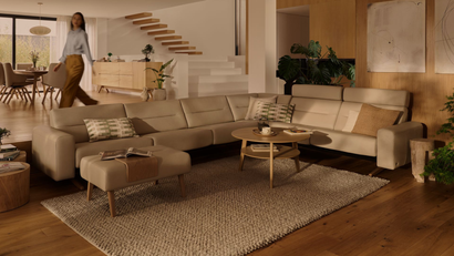 Warm living room with beige sectional sofa, wood accents, staircase, and a person walking in the background