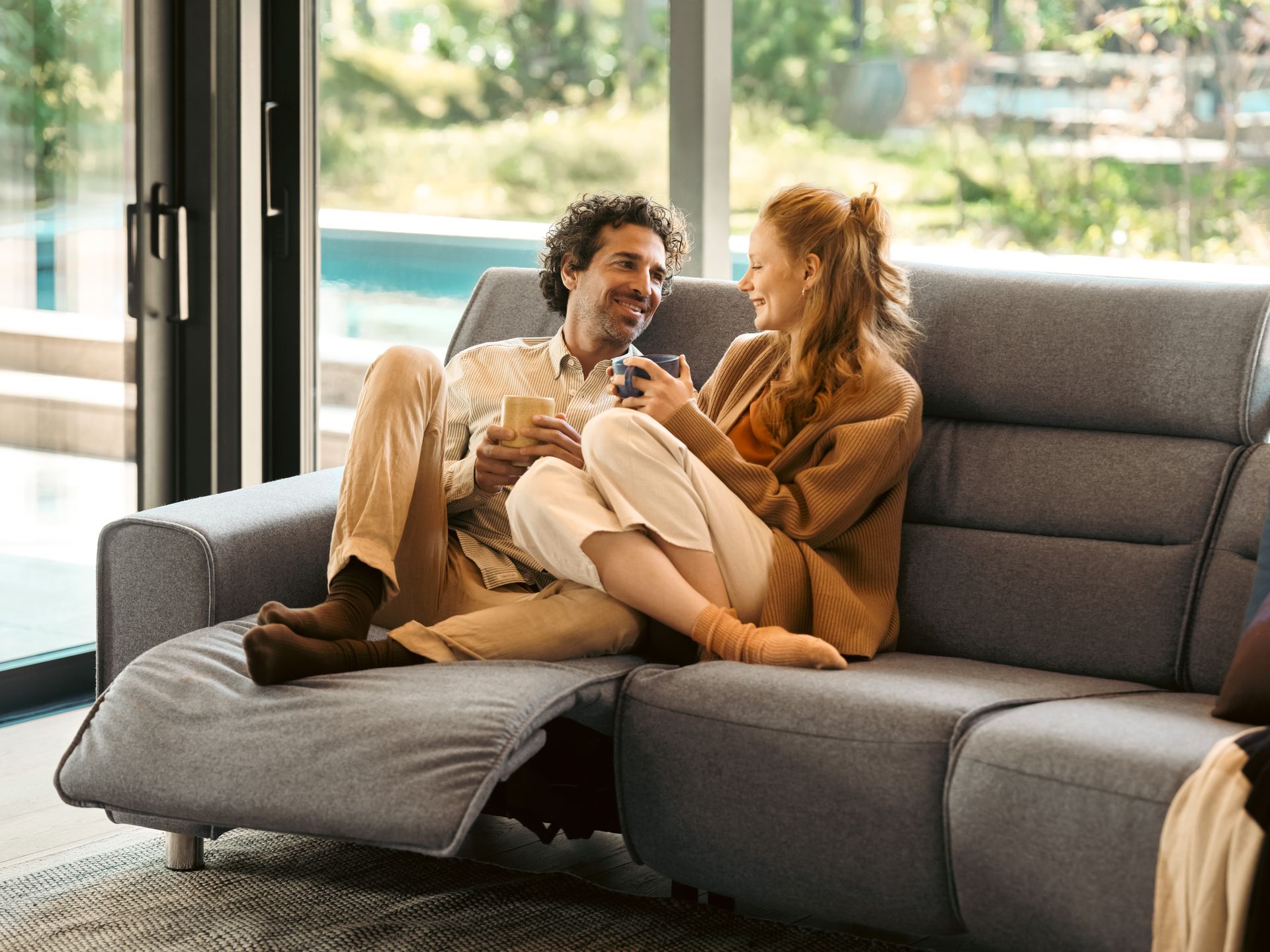 Person lounging on a gray couch with legs crossed on a wooden armrest, wearing gray pants and white socks.