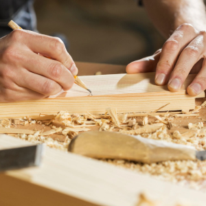 Hands carving a wooden piece with a chisel, surrounded by wood shavings in a workshop