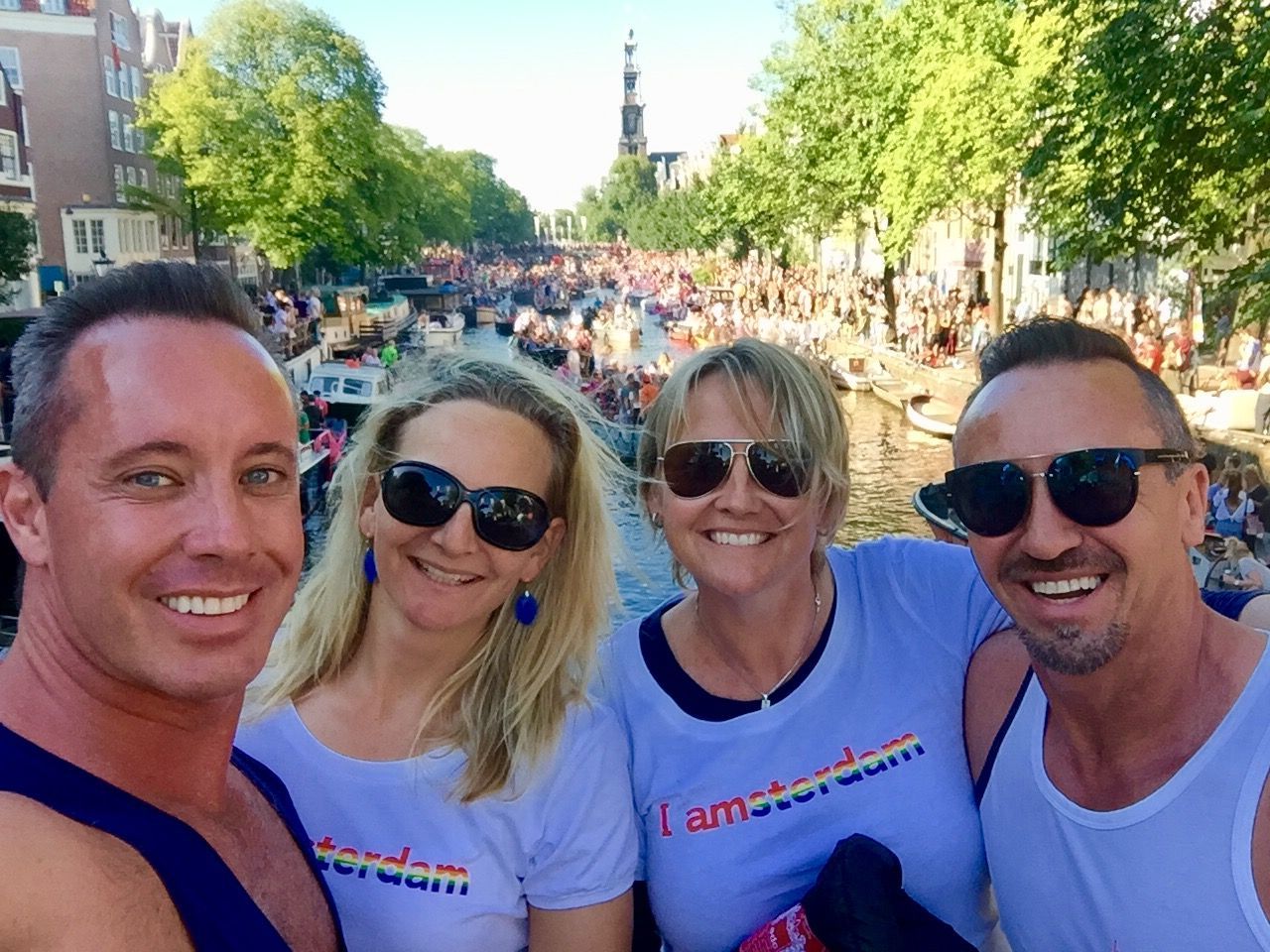Four people smiling for a selfie at a crowded outdoor canal festival on a sunny day