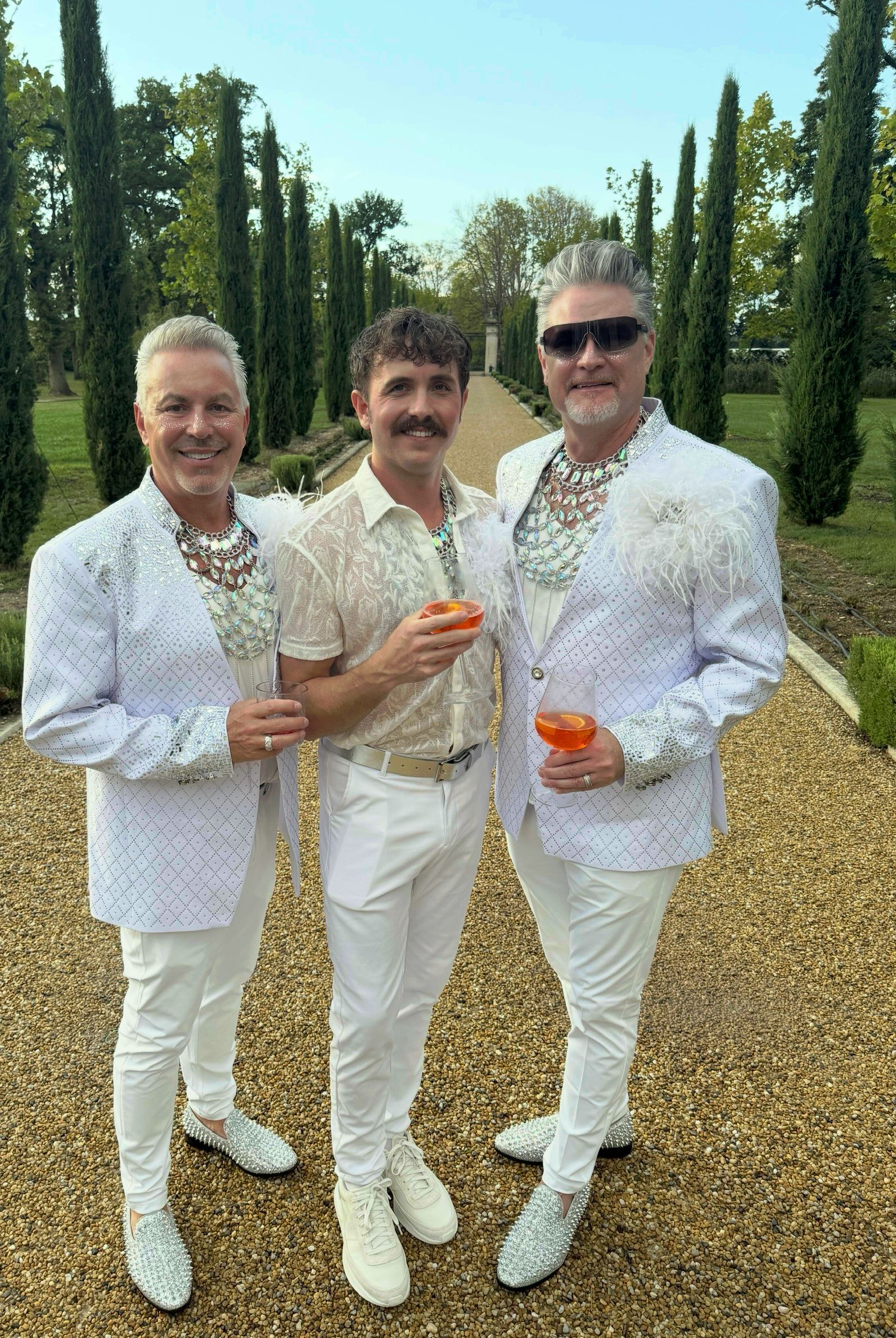 Three people in white suits holding orange drinks on a gravel path in a formal garden