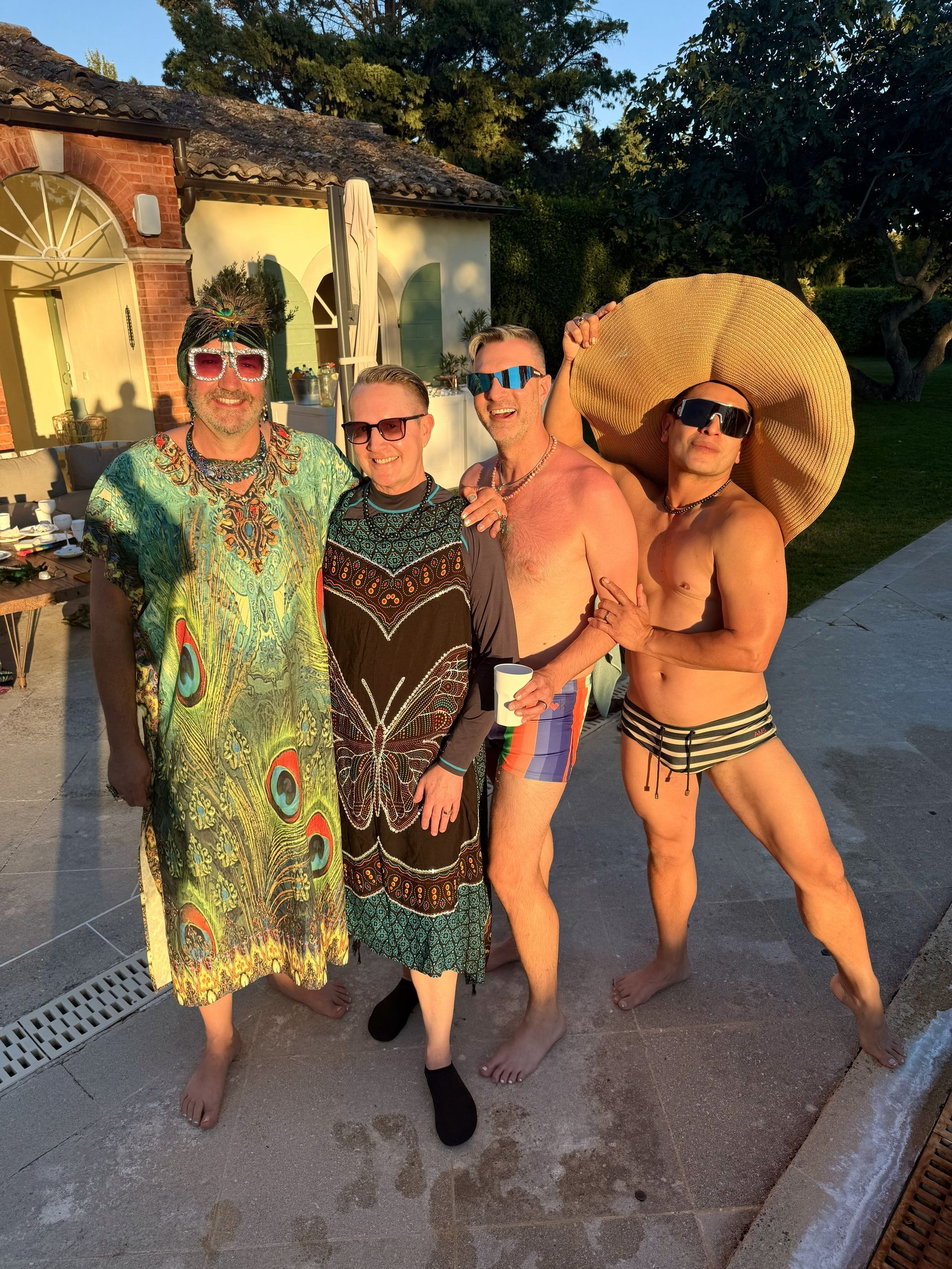 Four people in swimsuits and sunglasses posing outdoors at sunset by a pool.