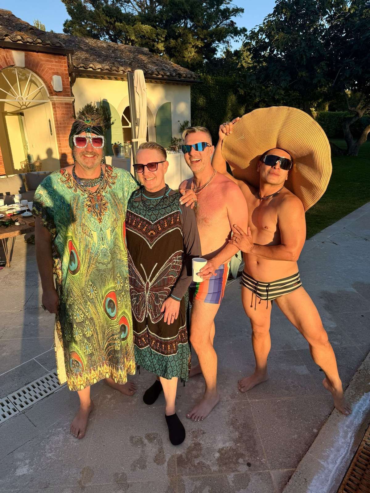 Four people in swimsuits and sunglasses posing outdoors at sunset by a pool.