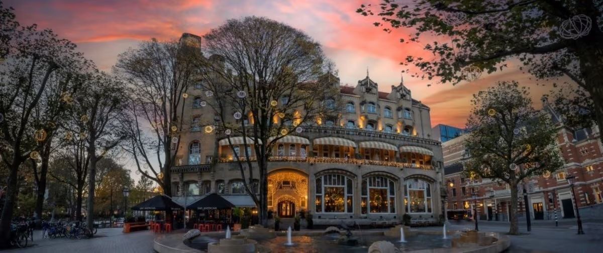 Historic building lit at dusk beneath a pink-orange sky, framed by leafless trees and parked cars.