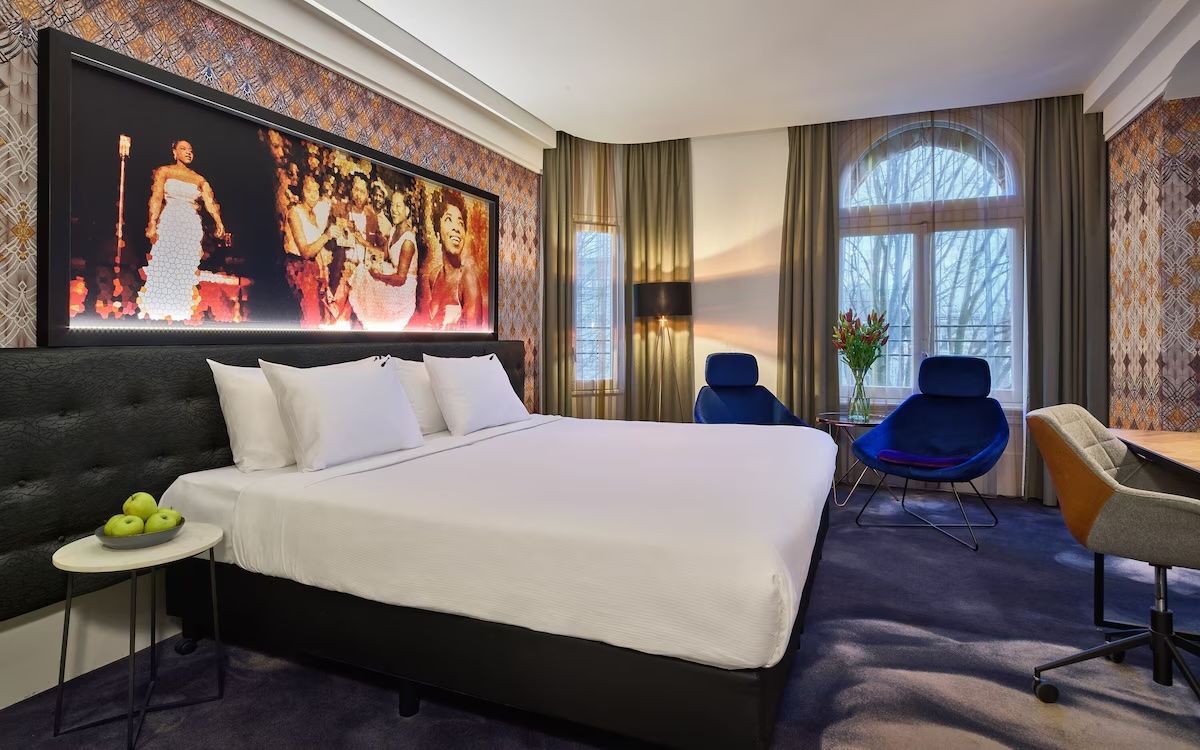 Modern hotel room with a king bed, city artwork, blue chair, and large window with curtains