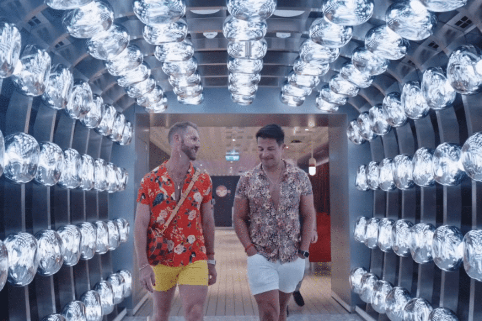 Two men in colorful shirts and shorts walking through a mirrored hallway with rows of hanging glass lights