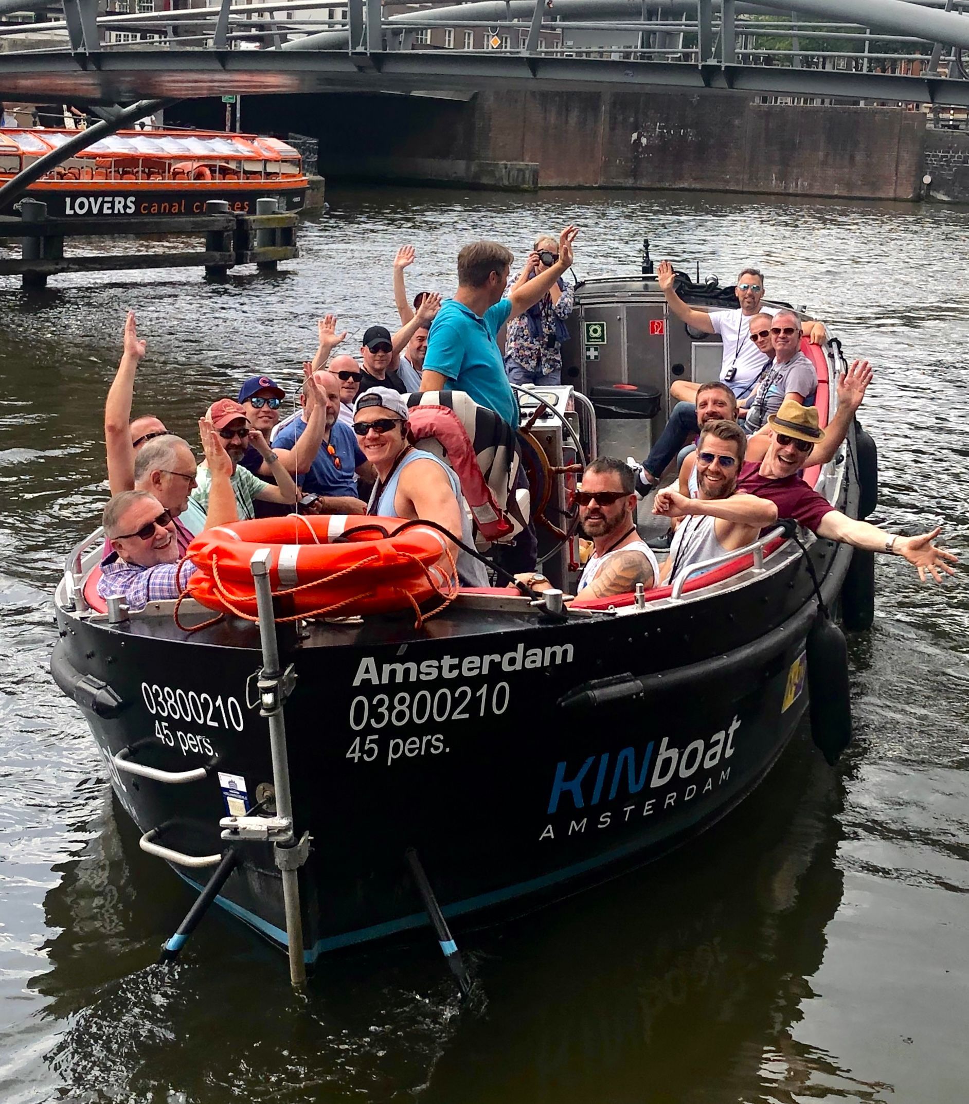 Tour boat with smiling passengers and raised arms on an Amsterdam canal under a bridge