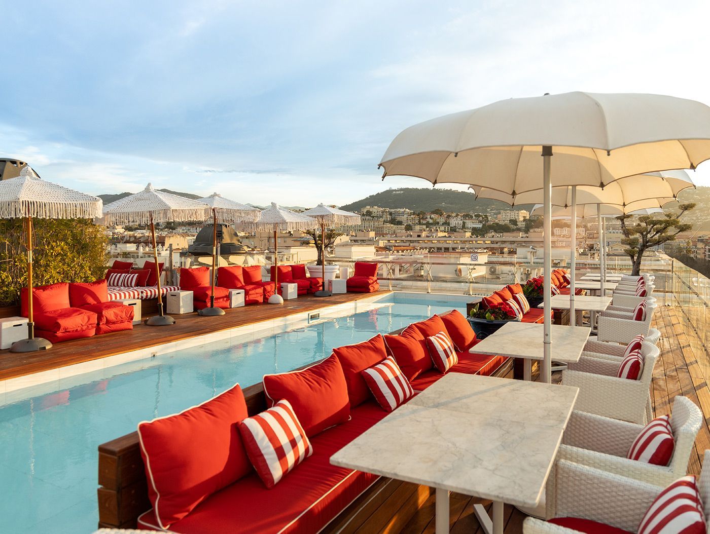 Rooftop lounge with red seating, tables, umbrellas, and a pool overlooking the city at sunset